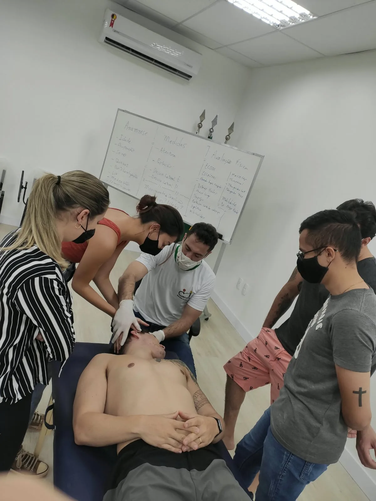 Aula prática em dupla, com o quadro de anamnese, medidas e avaliação física ao fundo